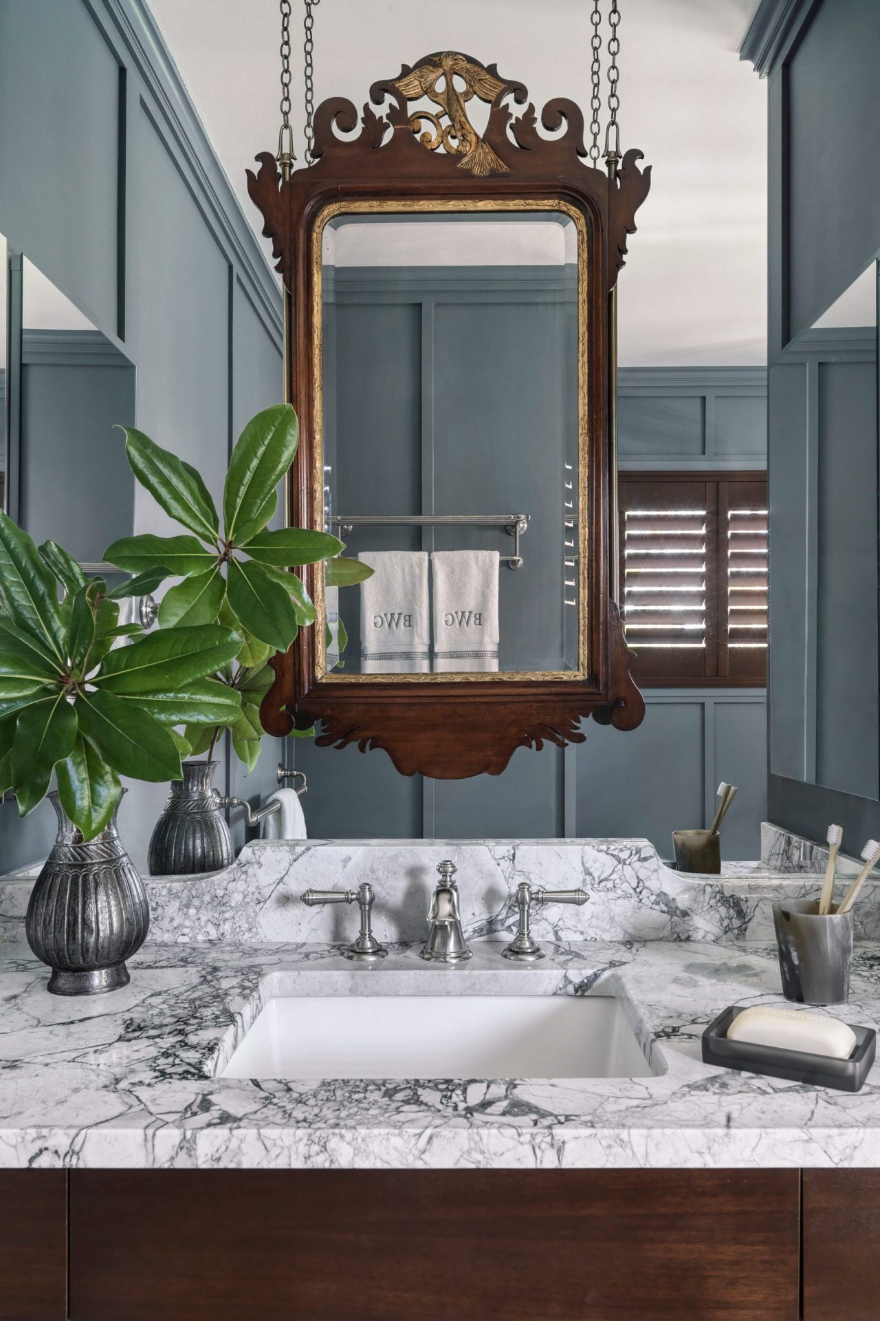 Blue-grey paneled wall with marble vanity, suspended carved wood-framed mirror, and elegant accessories like metal vases and soap trays.