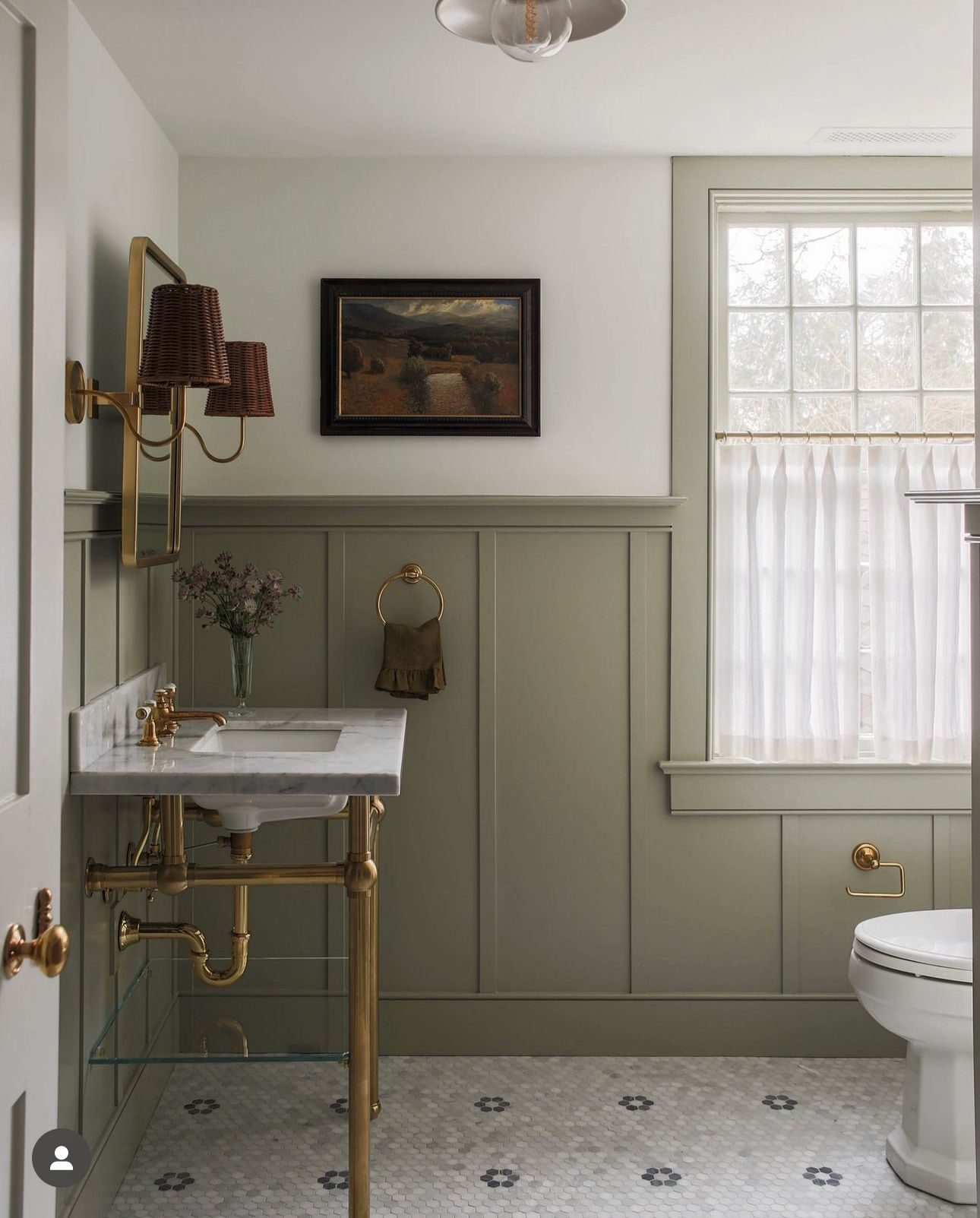 Sage green paneling, vintage artwork, brass basin stand, and white hexagonal tiles compose a soft, calming vintage-style bathroom.

