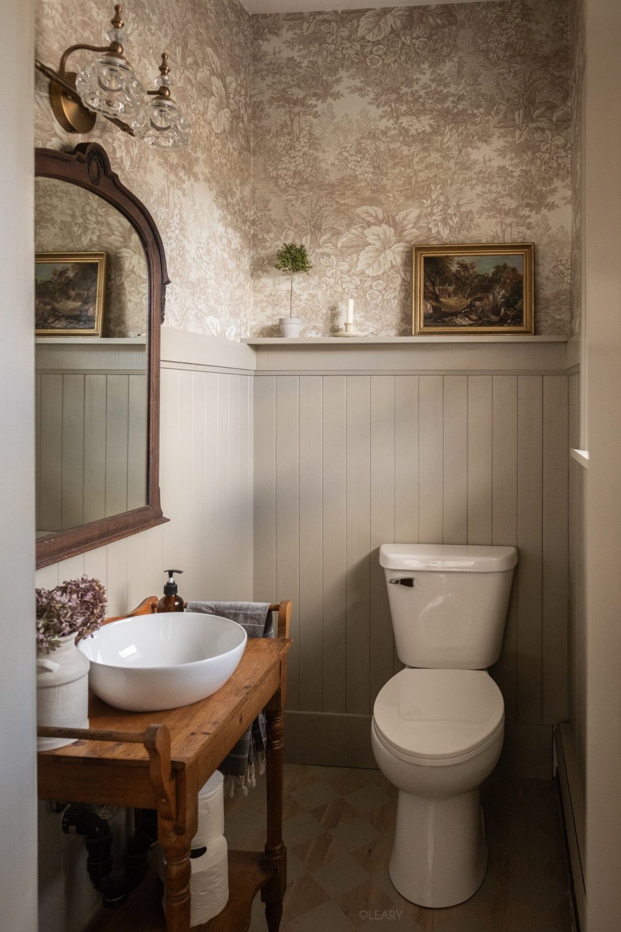 Vintage-inspired compact bathroom featuring a white ceramic countertop basin, classic wooden vanity, floral wallpaper, and warm decorative elements.