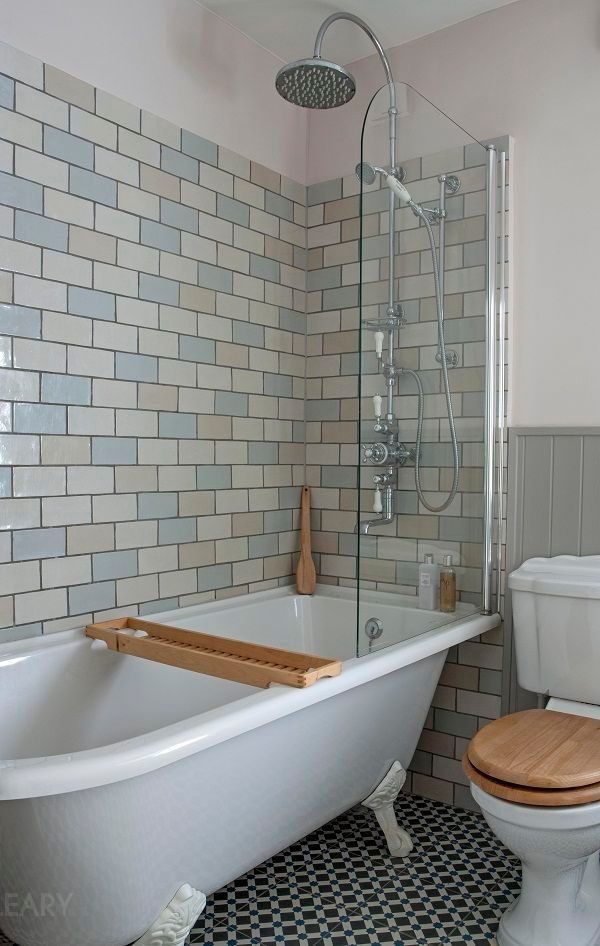 A compact bathroom featuring a white freestanding bathtub with clawfoot design, rainfall shower, glass partition, and vintage geometric tile flooring.