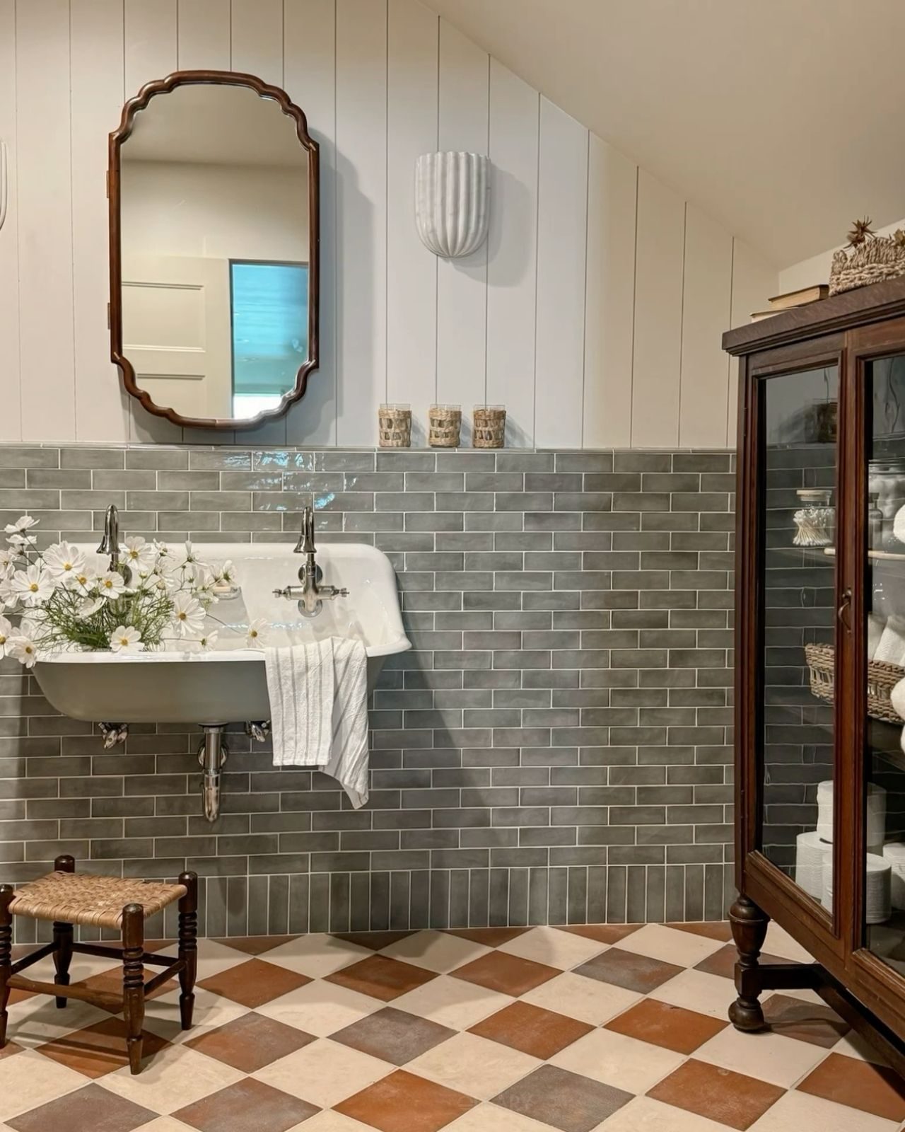 Country-style bathroom with vintage gray wall tiles, antique bathroom cabinet, built-in ceramic sink, wooden framed mirror, and a small woven stool.

