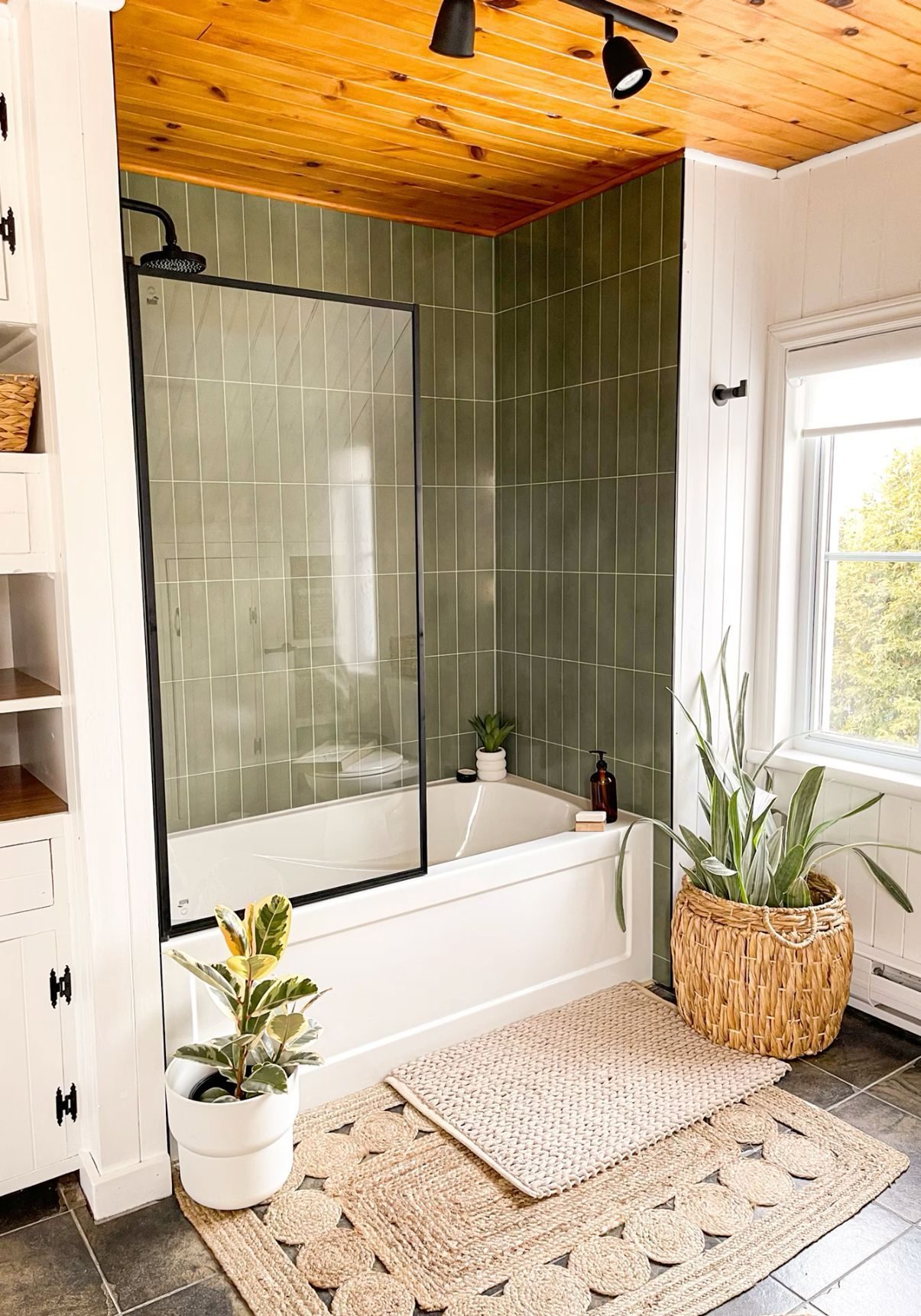 Modern minimalist bathroom with green tiles, a white bathtub, and a warm wooden ceiling, creating a cozy and well-lit space.

