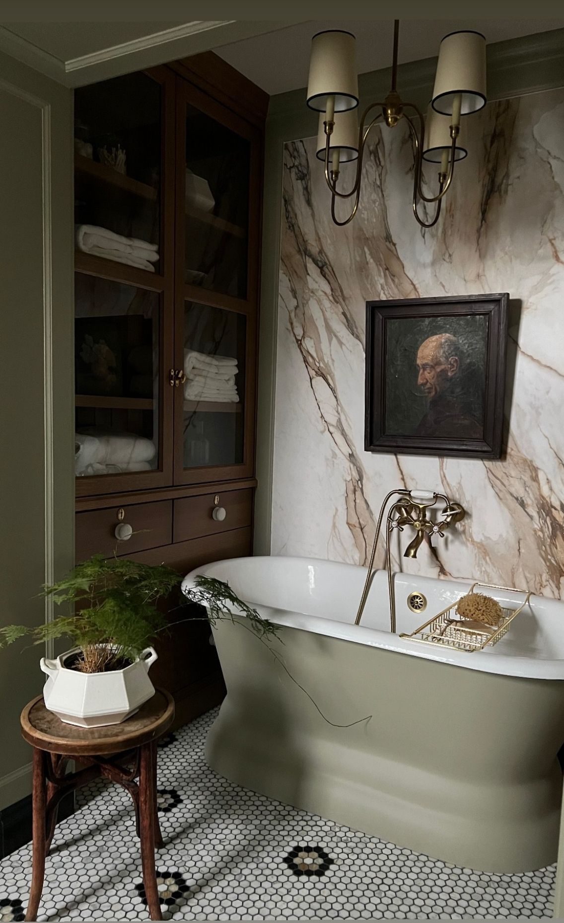 Sage-green pedestal bathtub with brass telephone faucet set against Calacatta-gold marble slab, vintage wood cabinet and hex-flower mosaic floor