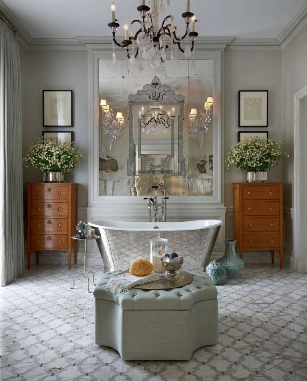 Hammered-nickel freestanding bathtub before antique mirror wall, crystal chandelier above, oak chests and arabesque marble mosaic floor.