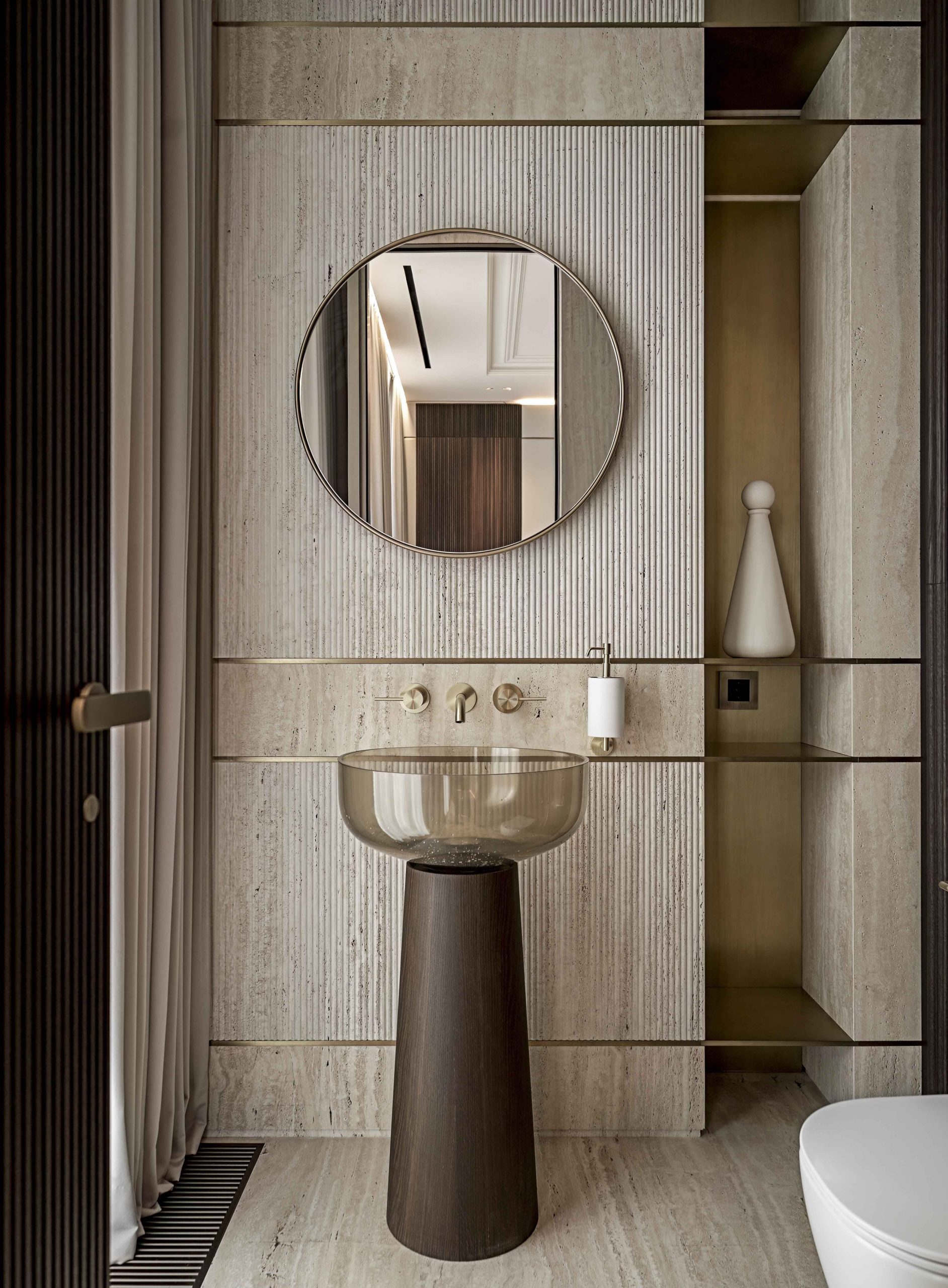 An elegant Joanna Laven-designed bathroom featuring a transparent, tinted resin basin on a dark wood pedestal, set against a fluted travertine wall with brass inlays.