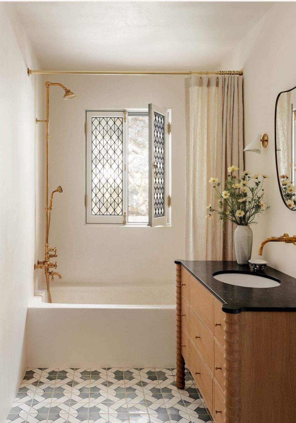 A modern Mediterranean-style bathroom featuring cream-colored walls, golden fixtures, patterned tile flooring, and a black stone vanity, lit with soft natural light.

