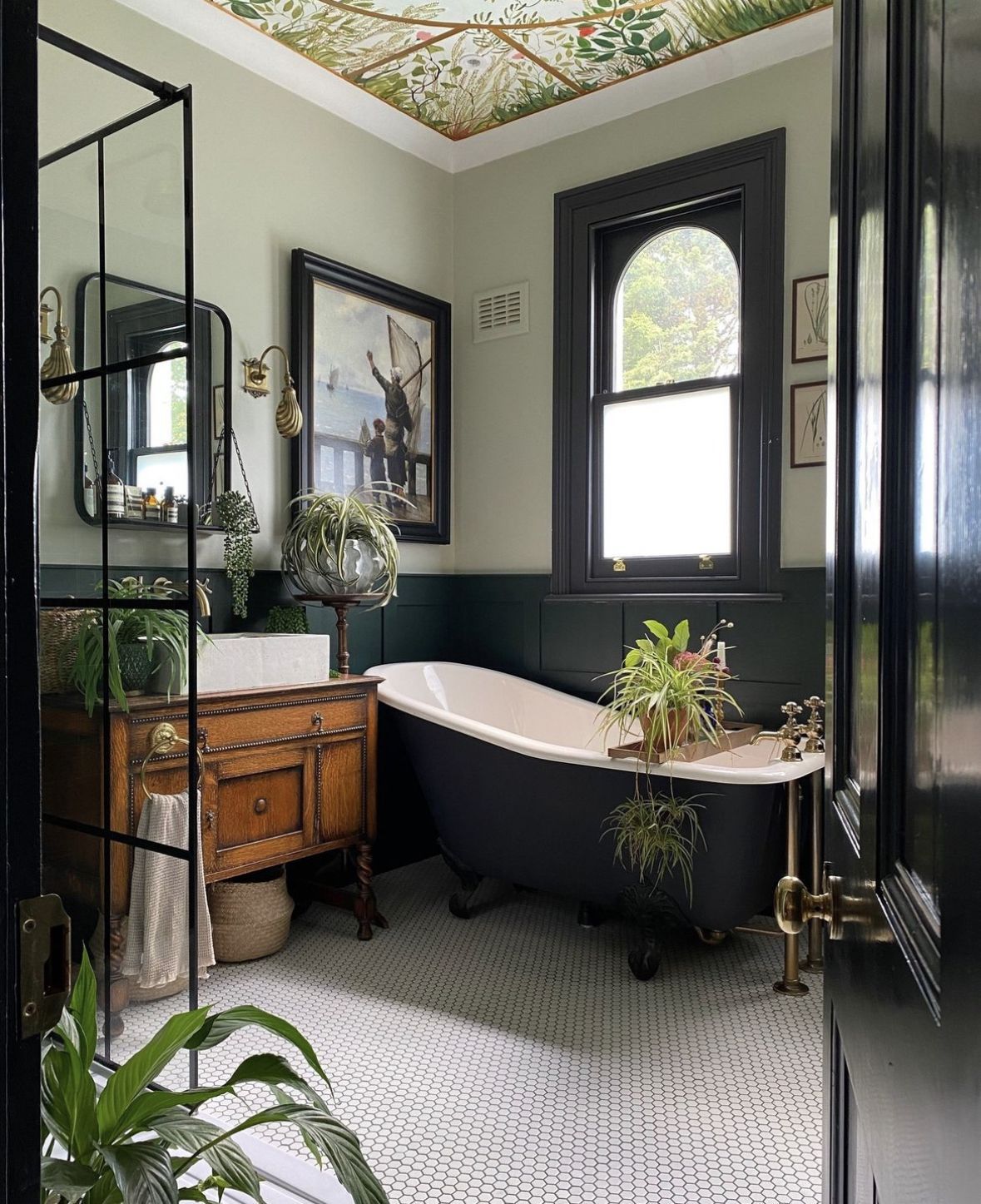 Vintage-inspired bathroom featuring a black claw-foot solid-surface bathtub, reclaimed-wood vanity with stone basin, botanical ceiling mural, brass fixtures and lush greenery.