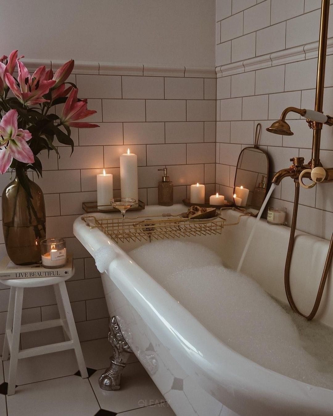 White solid-surface claw-foot bathtub with chrome feet, brass shower column, subway-tile walls, candles, pink lilies, and foam bubbles creating a spa ambience.