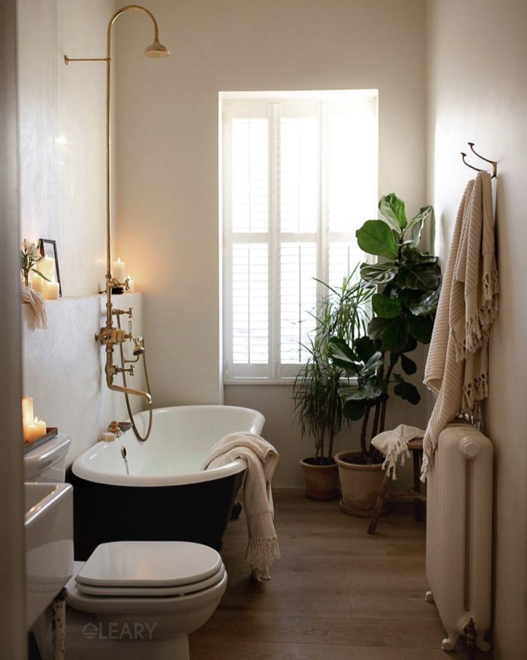 Narrow bathroom featuring a matte-black, white-interior slipper bathtub, exposed brass shower column, candles, wood floor and potted plants under shuttered daylight.