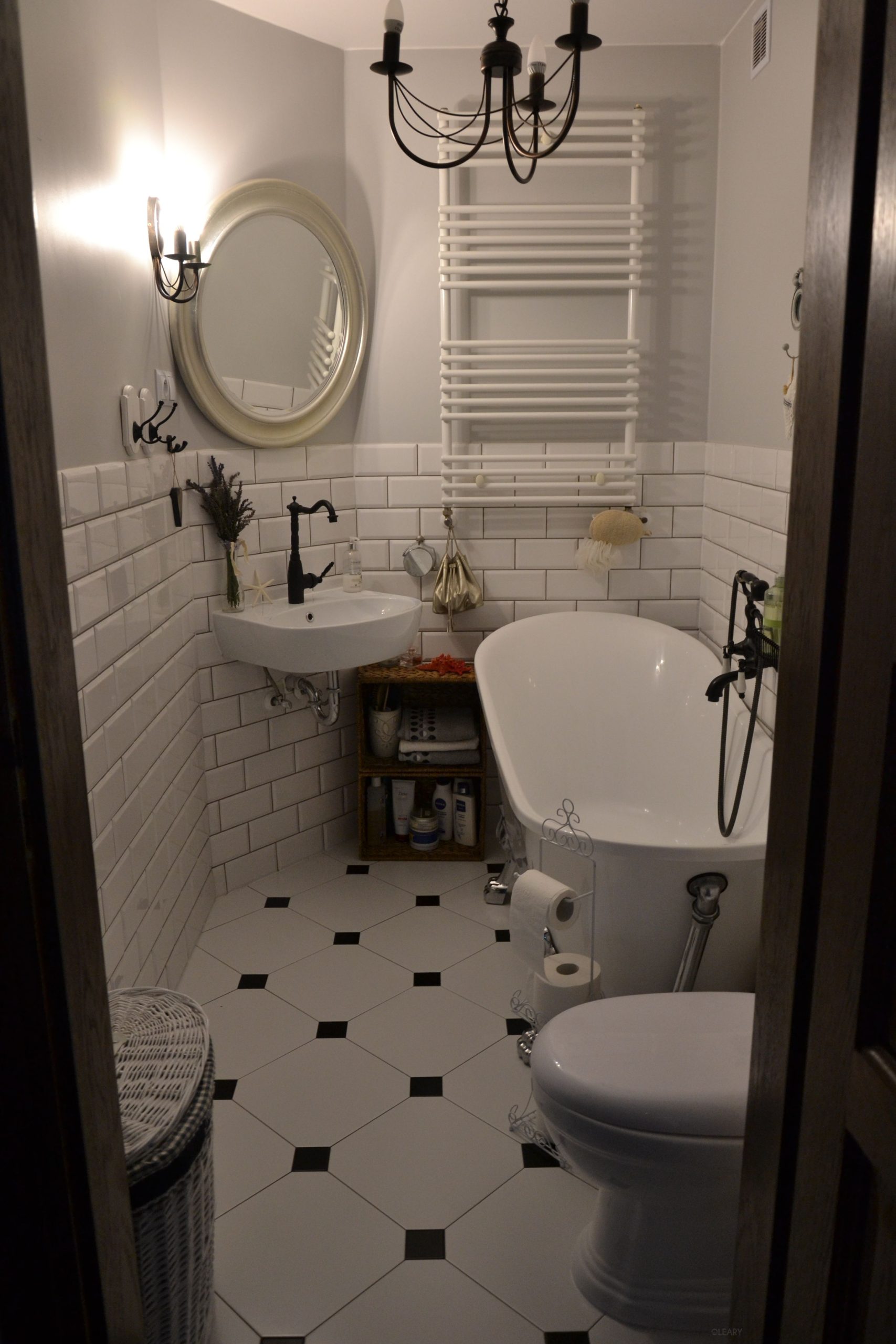 Narrow vintage bathroom with white solid-surface slipper bathtub on chrome legs, wall-hung semicircle basin, matte-black faucet and shower set, subway-tile walls, octagon-dot floor and chandelier sconce lighting.