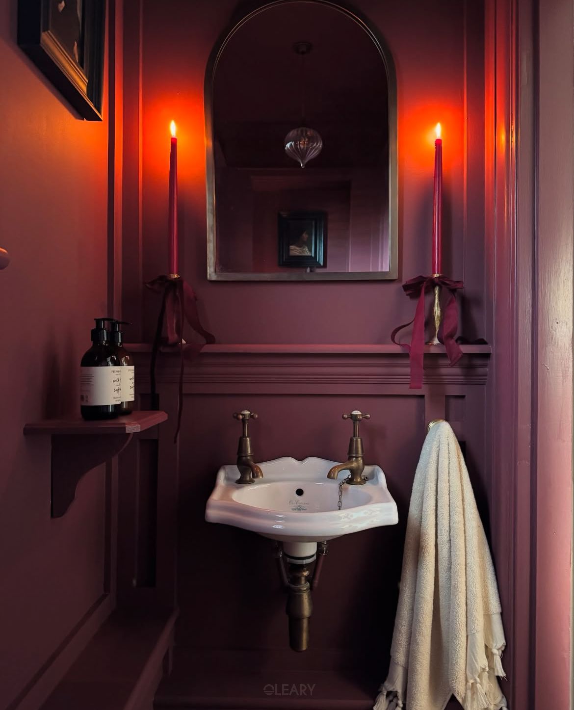Dark floral bathroom vignette featuring a gold-framed rounded mirror, globe-brass sconces, white marble countertop with integrated solid-surface basin, brushed-brass three-hole faucet and fresh flower arrangement.