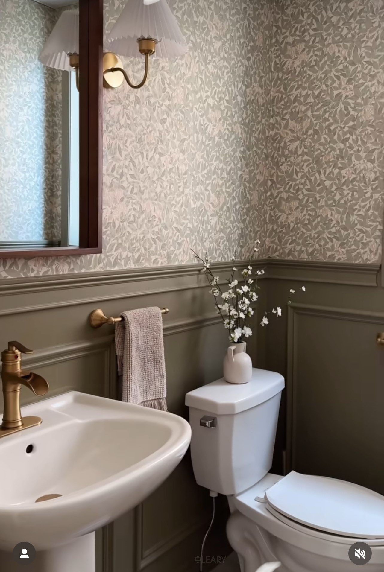 Cottage-style powder room with sage bead-mould wainscot, muted botanical wallpaper, white solid-surface pedestal sink, brushed-brass faucet and towel bar, pleated-shade wall sconce and white toilet topped with a ceramic bud vase.