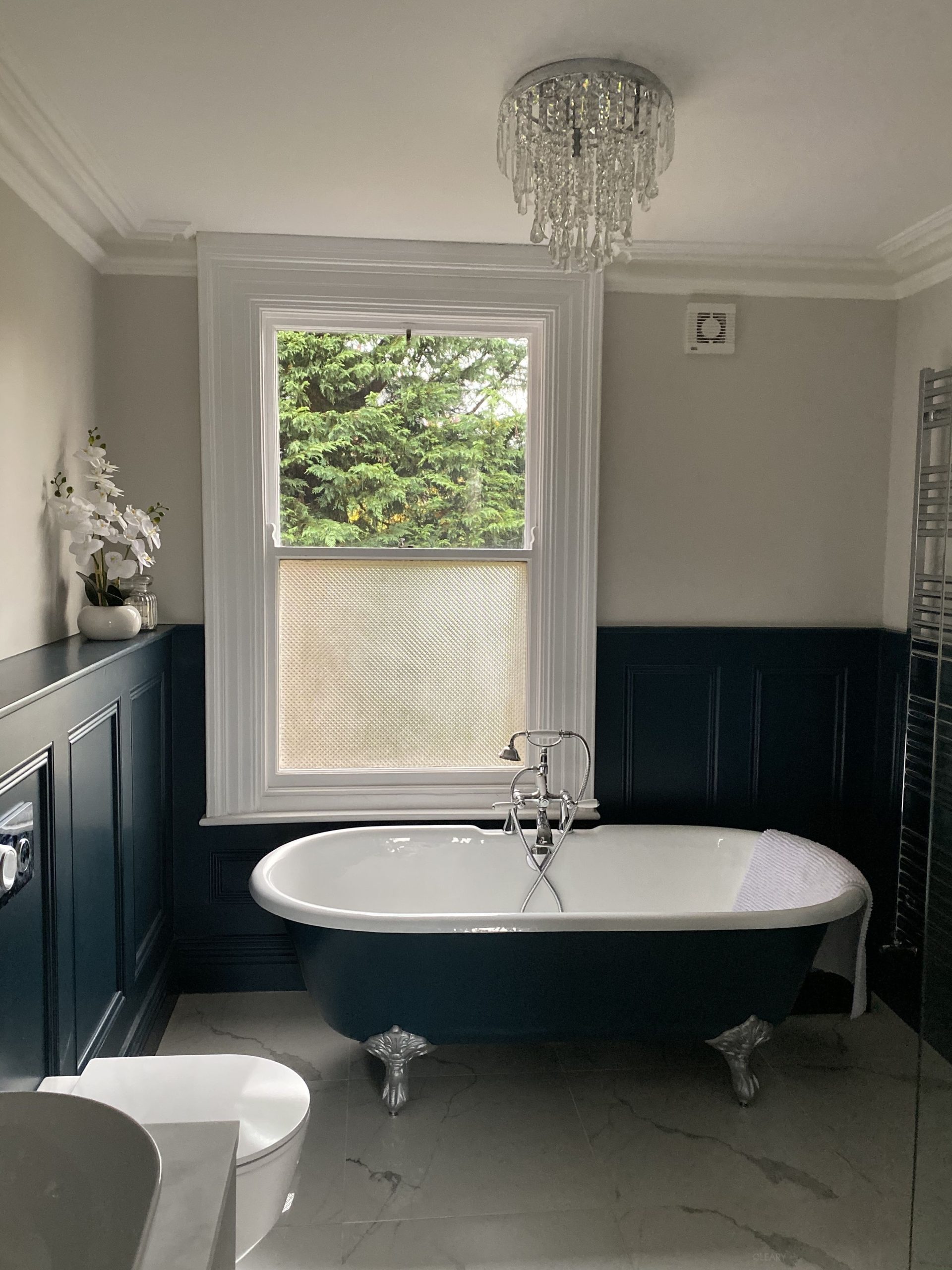 Modern-classic bathroom with navy blue claw-foot solid-surface bathtub, silver lion-paw feet, deep-panel wainscot, chrome telephone mixer, marble-look porcelain floor and crystal chandelier.