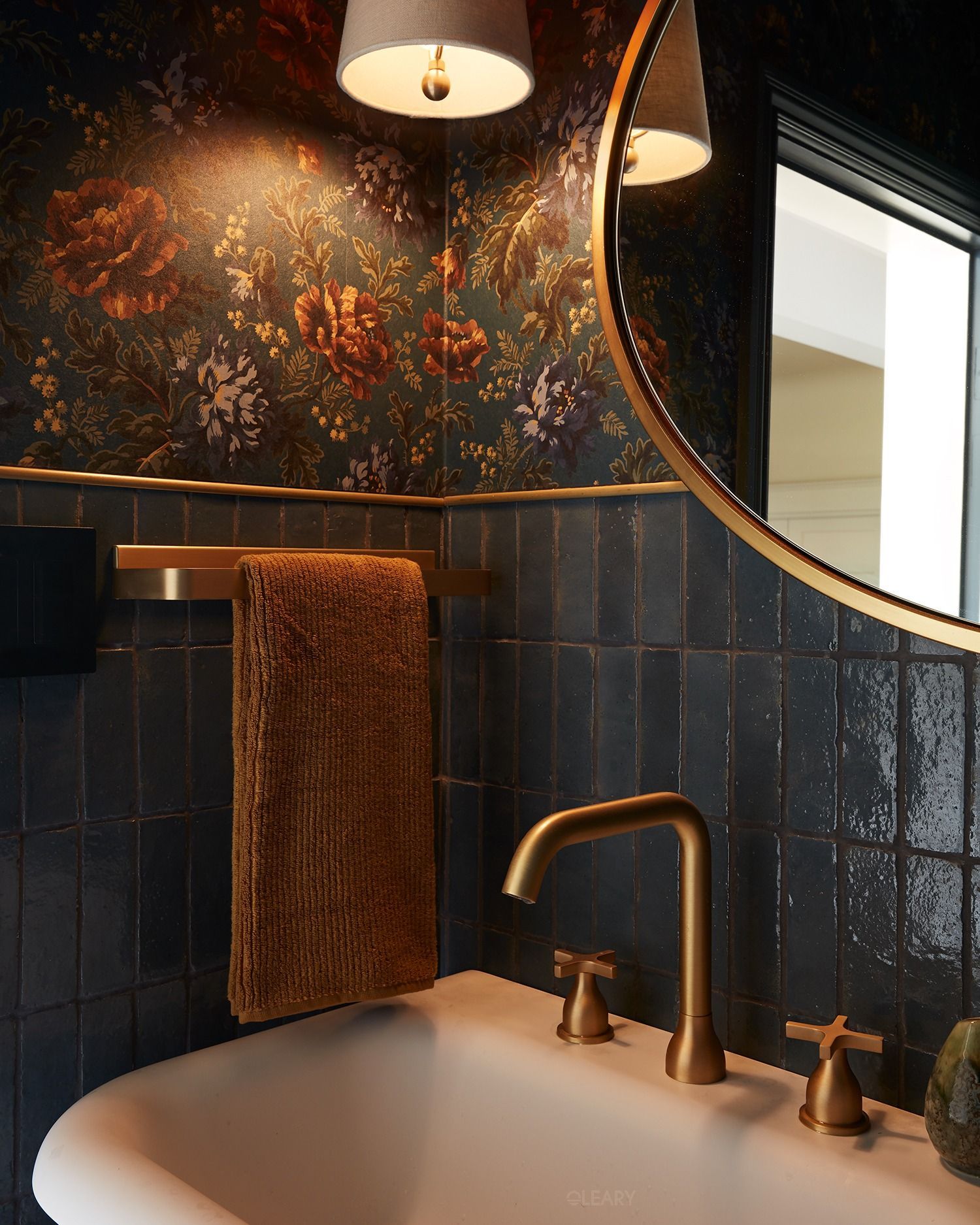 Close-up of a white solid-surface basin with brushed-brass deck-mount faucet set against midnight blue handmade tiles, brass towel rail, rounded brass mirror and dark floral wallpaper.