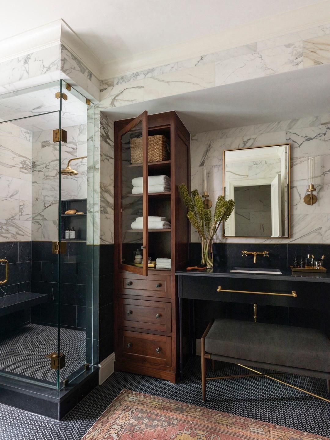 A luxurious modern bathroom featuring white marble walls and dark black tiles, accented with gold fixtures. It includes a glass-enclosed walk-in shower with a built-in black shower bench and niche. A dark wood tall cabinet stands beside a black vanity with a gold faucet and a gold-framed mirror. The floor is laid with dark mosaic tiles.