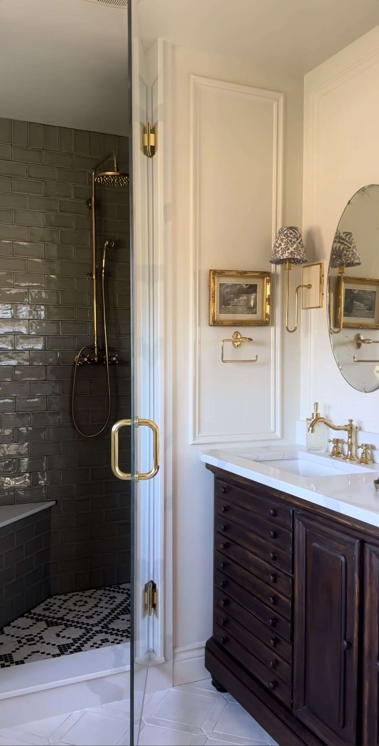 A classic luxury bathroom, reflecting 2025 design trends, featuring a dark-tiled walk-in shower with brass fittings (including a built-in shower bench and mosaic floor tiles), a dark solid wood multi-drawer vanity with a white stone countertop and undermount basin, vintage brass faucets, an oval bathroom mirror, and elegant wall sconces, with exquisite paneling on the walls.