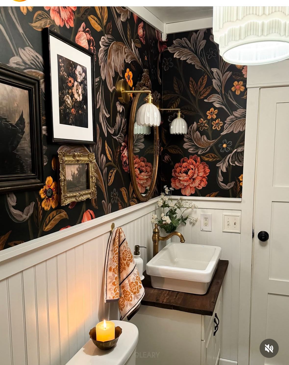 A vintage artistic style powder room, reflecting 2025 design trends, featuring a dark background large floral print wallpaper contrasting with white vertical wainscoting. A solid wood countertop holds a white square ceramic countertop basin, complemented by a vintage brass wall-mounted faucet. An oval gilt-framed mirror is positioned above, with a double-arm vintage wall sconce.