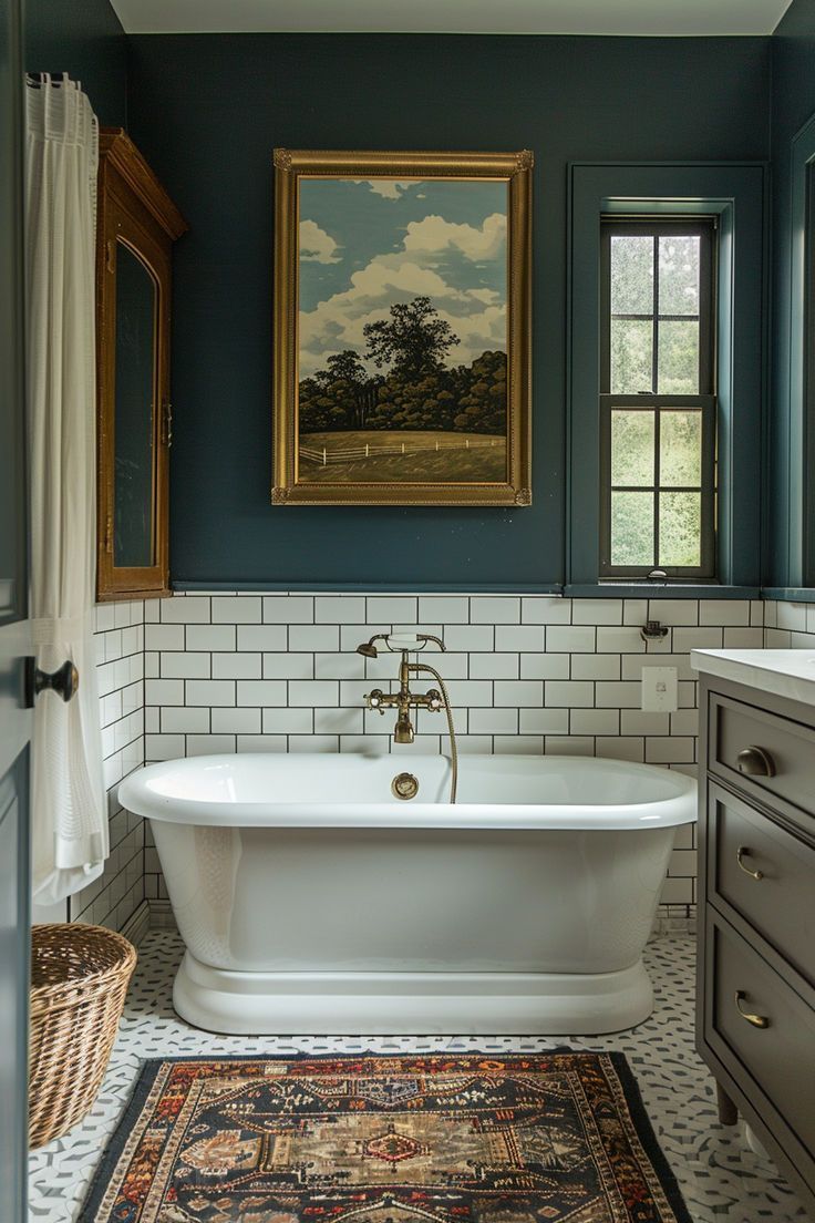 A vintage eclectic style bathroom, reflecting 2025 design trends, featuring a white freestanding pedestal bathtub with a brass tub filler. The upper wall is a deep teal/blue, with white subway tiles below. The floor combines vintage patterned tiles with a Persian-style rug. A grey custom vanity is on the right, and a recessed wooden glass-door cabinet is on the left wall. The window has a dark frame.