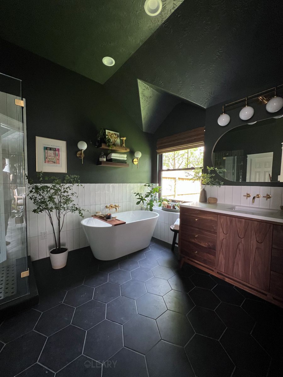 A luxurious 2025 moody contemporary bathroom featuring a white freestanding oval soaking tub, dark green or charcoal painted walls and angled ceiling, a custom walnut double vanity with a light stone countertop, gold/brass faucets and lighting fixtures, white vertical tile wainscoting, and dark hexagonal floor tiles.