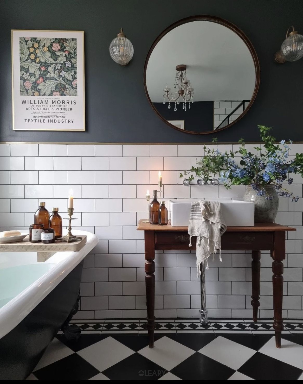 A 2025 modern vintage style bathroom featuring a white rectangular ceramic countertop basin on a rustic dark wood table vanity, against a backdrop of dark grey upper walls and white subway tile wainscoting. A large round dark-framed mirror is flanked by vintage industrial sconces, with a black and white patterned tile floor and a partially visible dark freestanding bathtub.