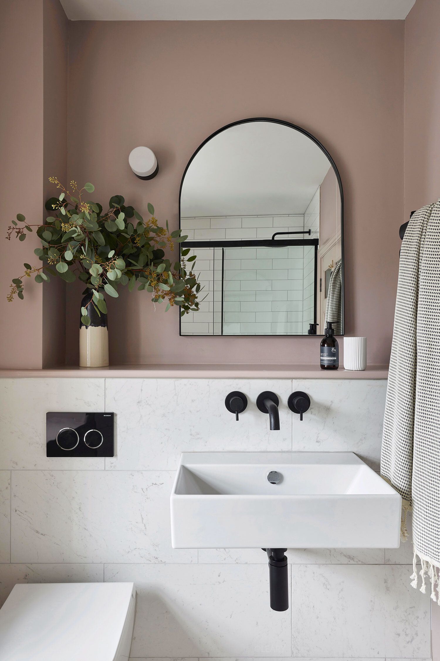 A 2025 modern minimalist bathroom featuring a white rectangular wall-hung ceramic basin with matte black wall-mounted faucets. An arched black-framed mirror hangs above, against a dusty rose painted upper wall. The lower wall has light marble-effect tile wainscoting with an integrated display ledge. A wall-hung toilet with a black flush plate is partially visible.