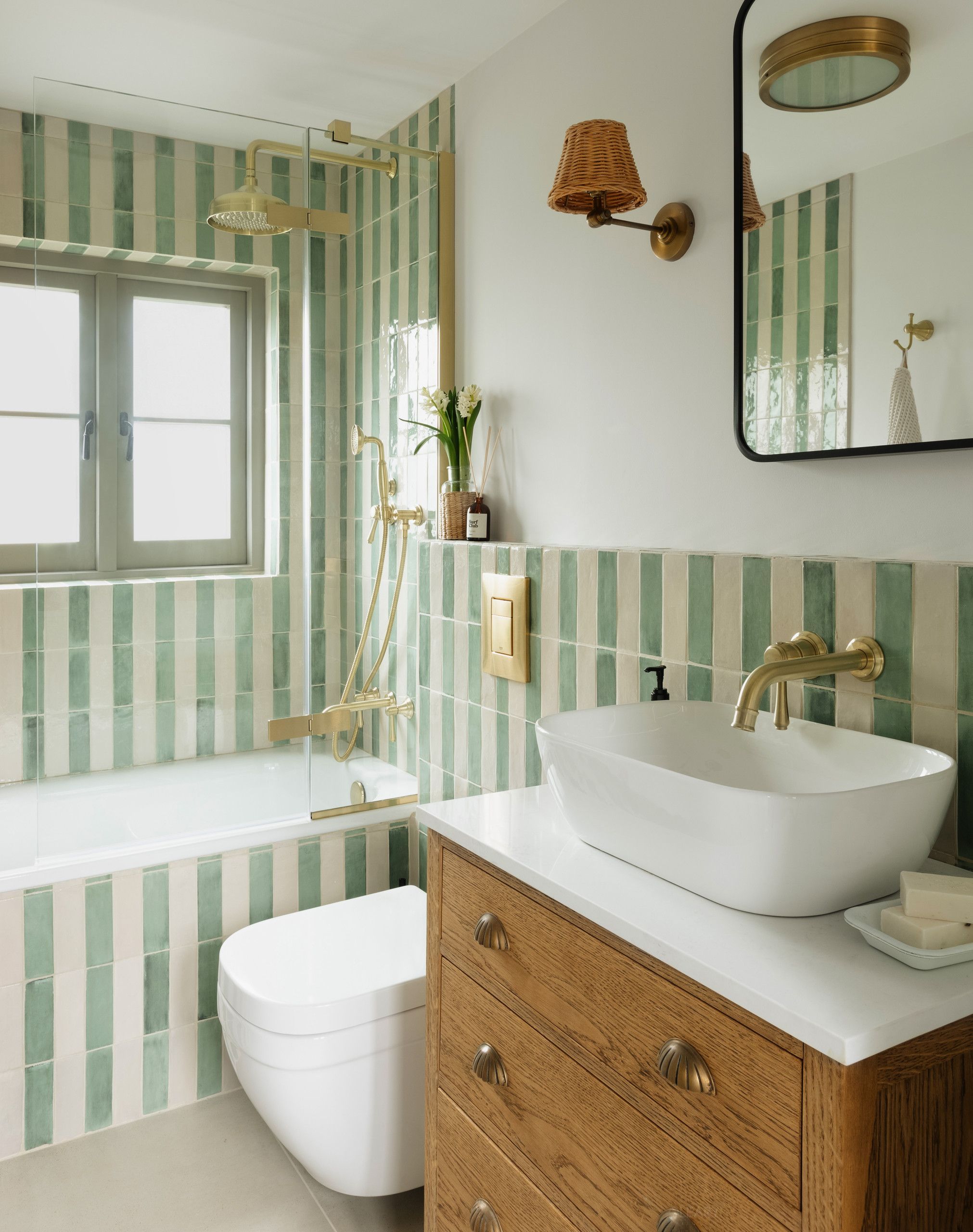 A 2025 modern classic bathroom design featuring a white organic-shaped ceramic countertop basin on a natural oak vanity with a white countertop. The walls showcase green and white vertical striped tiles extending to the built-in bathtub surround, complemented by brass/gold fixtures, a modern toilet, a rectangular mirror with rounded corners, and a woven rattan wall sconce.