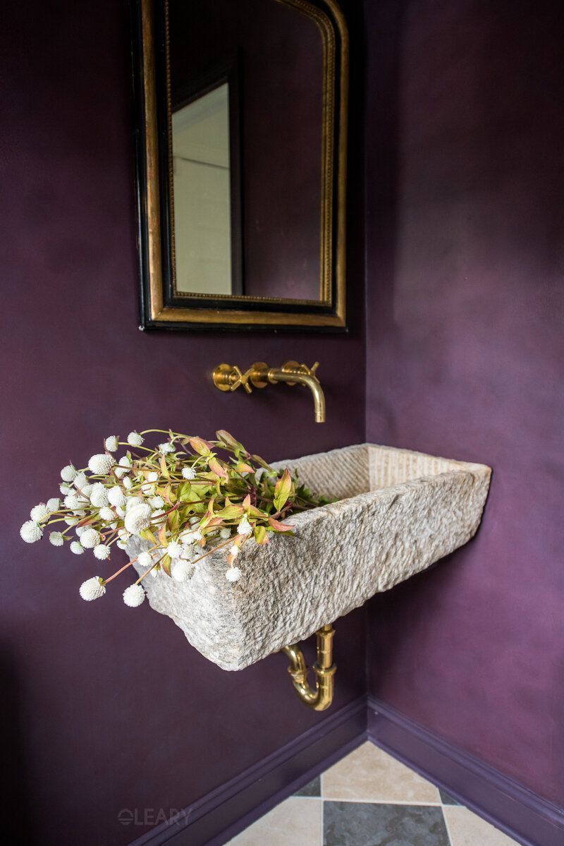 A luxurious 2025 rustic-luxe bathroom featuring a unique wall-hung rough-hewn natural stone basin (washbasin) with an antique brass wall-mounted faucet and exposed brass P-trap. An ornate dark wood and gilt framed mirror hangs above, set against a deep aubergine or plum-colored textured wall, with a classic black and white checkerboard tile floor.