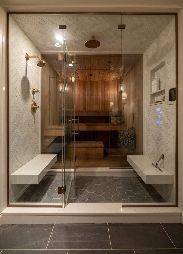A luxurious 2025 custom home spa featuring a large glass-enclosed steam room and shower. The steam room interior has natural wood paneling and benches. An antechamber within the enclosure showcases light herringbone tiled walls, white floating benches, recessed wall niches with glass shelves, traditional brass shower fixtures, and dark hexagonal floor tiles.