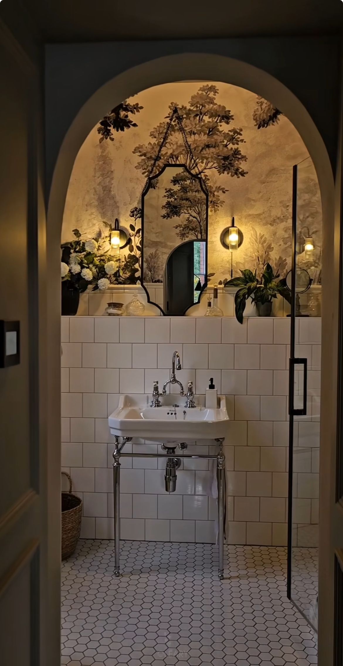 A 2025 Modern Farmhouse style bathroom featuring a custom sage green or grey-green painted wooden vanity with a white Carrara marble countertop, undermount sink, and a traditional brass bridge faucet. An ornate vintage gold framed rectangular mirror hangs above, against a soft grey textured wall, complemented by a classic wall sconce and elegant floral arrangements.
