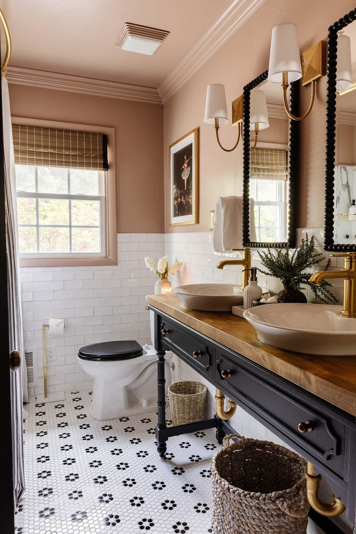 A 2025 eclectic vintage style bathroom featuring two beige ceramic countertop basins on a custom dark wood console vanity with a warm wood countertop and antique brass faucets. Ornate black-framed mirrors with gold accents hang above, flanked by vintage wall sconces, against a backdrop of soft pink upper walls and white subway tile wainscoting. The floor is a classic black and white floral patterned mosaic tile.