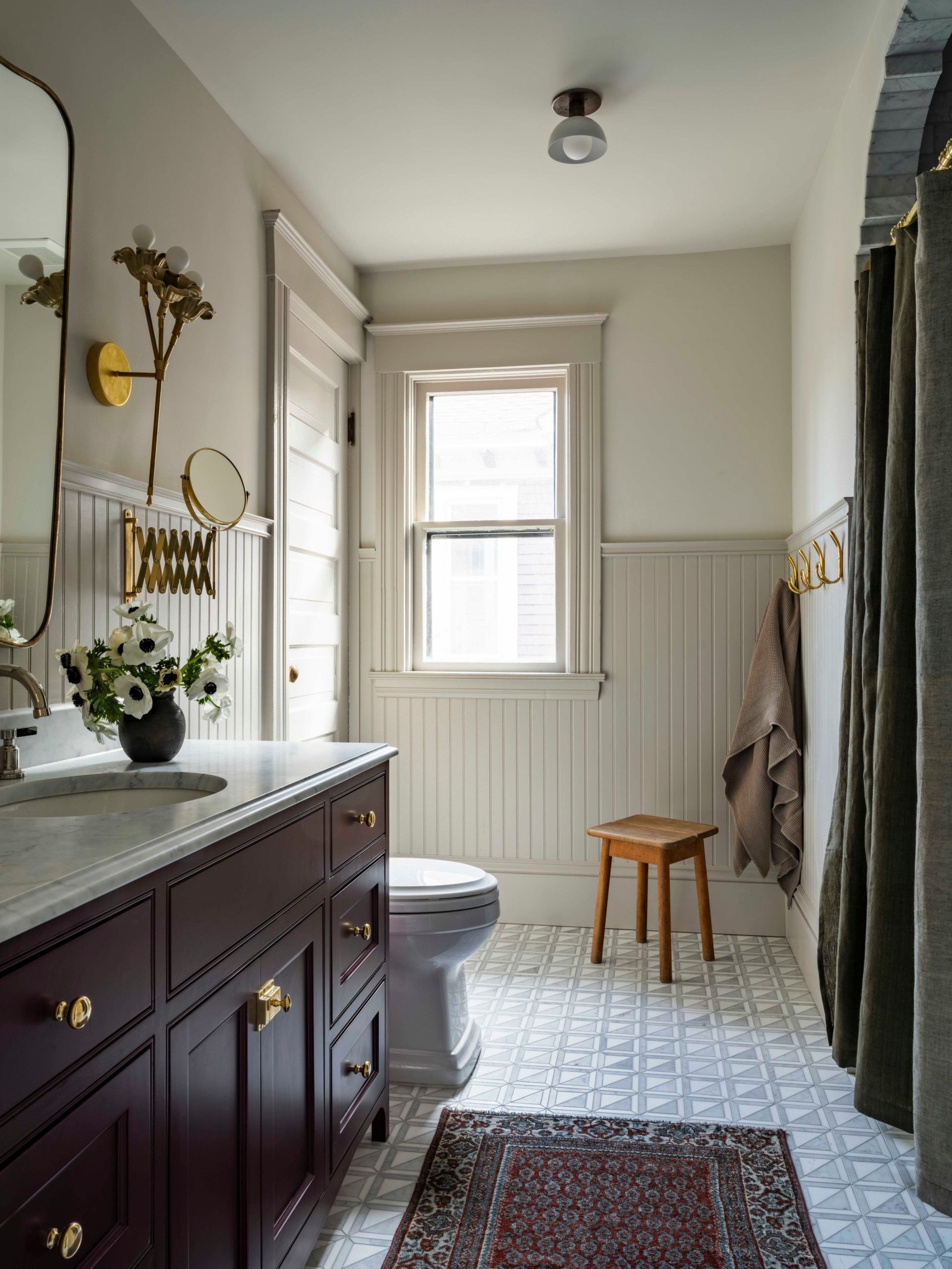 A luxurious 2025 classic traditional bathroom featuring a custom dark wood vanity with a white marble or engineered stone countertop and an undermount ceramic sink. Polished chrome widespread faucets with cross handles adorn the basin, complemented by a large rectangular mirror, traditional wall sconces, extensive light-colored classic wall paneling, and a patterned mosaic tile floor.

