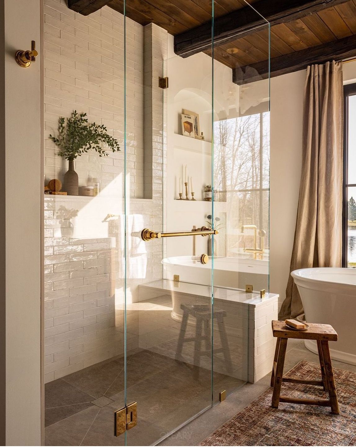 A luxurious 2025 rustic modern bathroom featuring a large, glass-enclosed shower room with light textured Zellige-style tiled walls, built-in recessed niches with glass shelves, and antique brass shower fixtures. A white freestanding bathtub is partially visible beyond the shower, under a window, complemented by dark wood ceiling beams and rustic wooden accessories.

