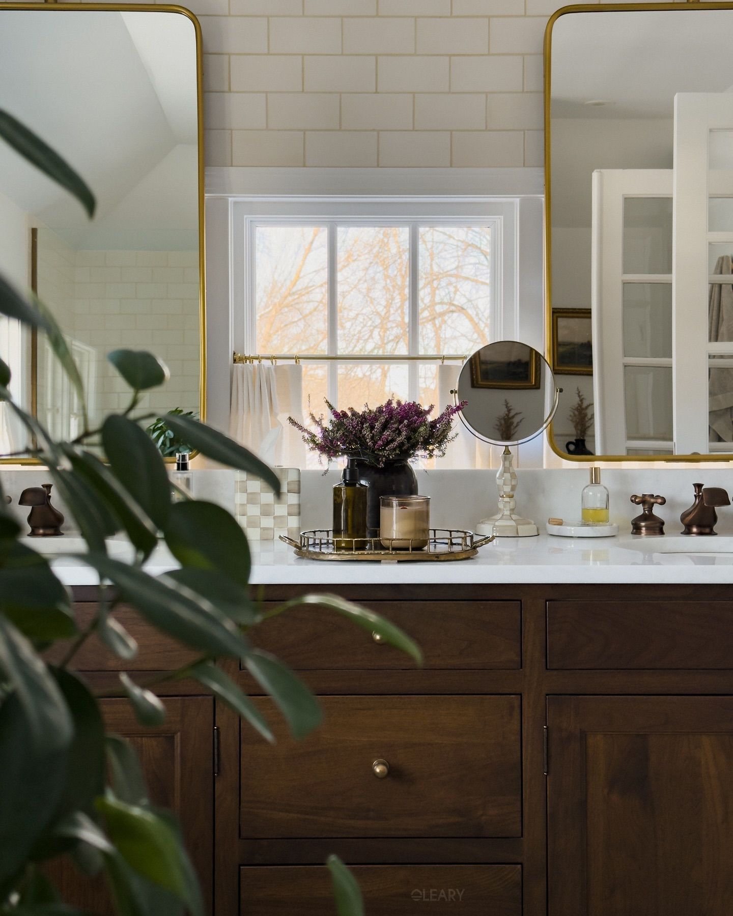 A luxurious 2025 classic traditional bathroom featuring a custom dark wood double vanity with a light marble or engineered stone countertop, two undermount white ceramic sinks, and traditional brass/gold widespread faucets. Twin large rectangular mirrors with ornate gilt frames hang above, flanked by classic double-arm wall sconces with fabric shades, against light neutral walls with detailed crown molding.

