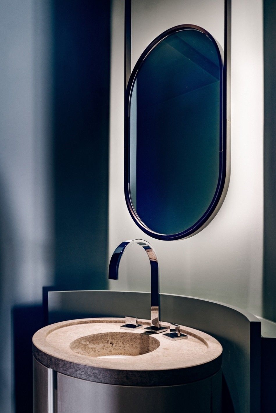 Ultra-modern luxury bathroom with a light natural stone cylindrical pedestal sink, a polished chrome faucet, a deep blue layered curved wall with hidden backlighting, and a dark-framed oval mirror.
