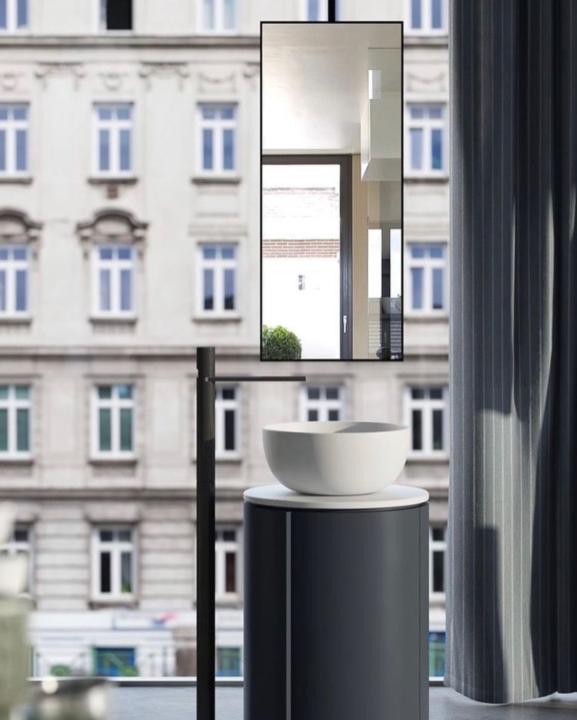 Cylindrical pedestal vanity, white bowl sink, black floor-mounted faucet, suspended rectangular mirror, urban bathroom design, modern minimalist, dark grey vanity, compact bathroom solutions, contemporary vanity, minimalist bathroom, city apartment bathroom, boutique hotel bathroom, sleek fixtures, space-saving design, refined interior, tall mirror, wall-to-wall window, matte black faucet