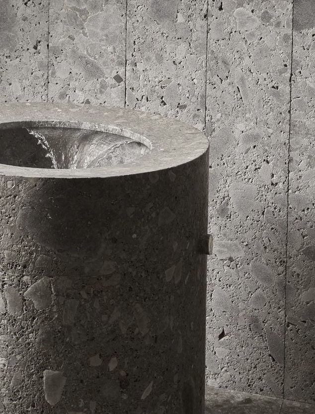 Close-up of a dark grey terrazzo or concrete cylindrical freestanding pedestal sink with water flowing from its center, against a light grey terrazzo wall, highlighting raw material beauty.