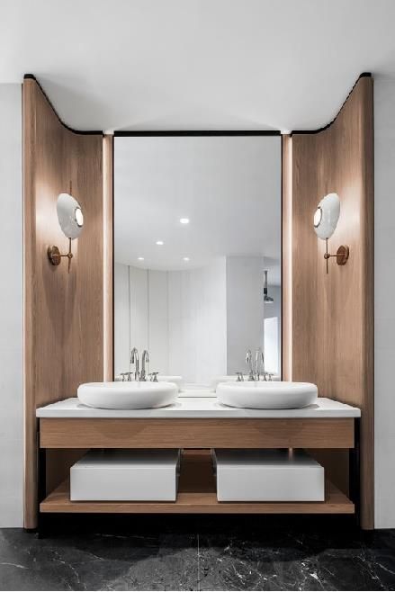 Modern luxury double bathroom vanity set within a light wood curved alcove with a large mirror, two white bowl sinks, and unique oval wall sconces, on a dark marble floor.