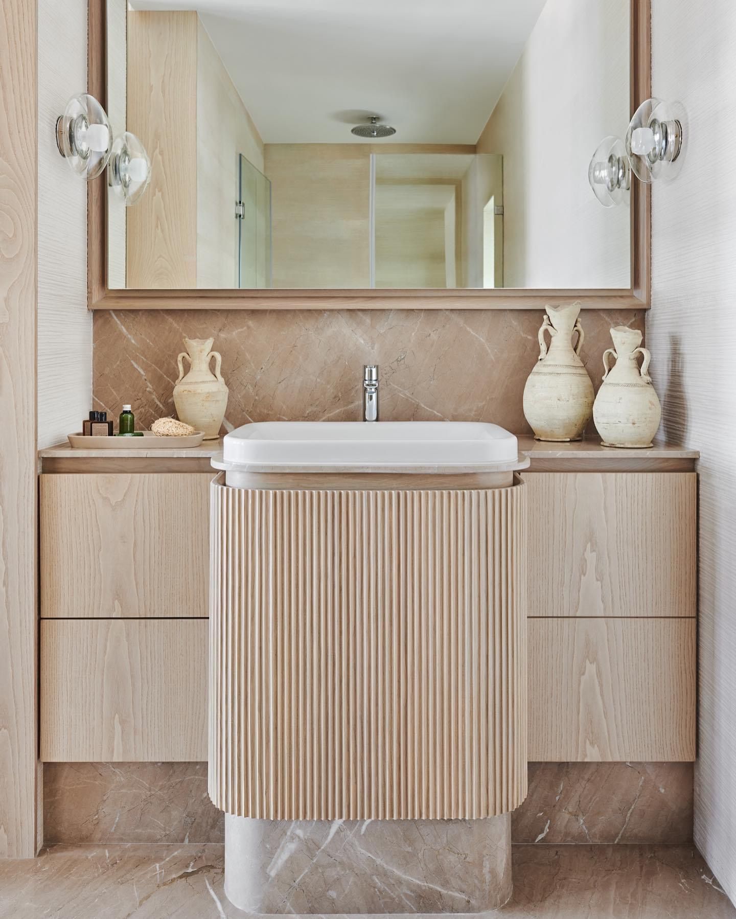 Modern bathroom featuring a light wood fluted vanity with a white integrated sink, a brown marble backsplash, and a large rectangular mirror, flanked by unique glass wall sconces.