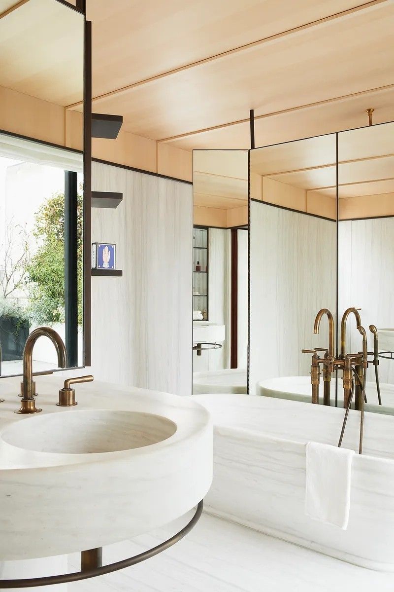 A luxurious modern bathroom featuring a white marble circular washbasin and a matching freestanding marble bathtub. The space is accented with vintage bronze faucets, a large window overlooking greenery, and a sophisticated blend of white stone and warm wood paneling.