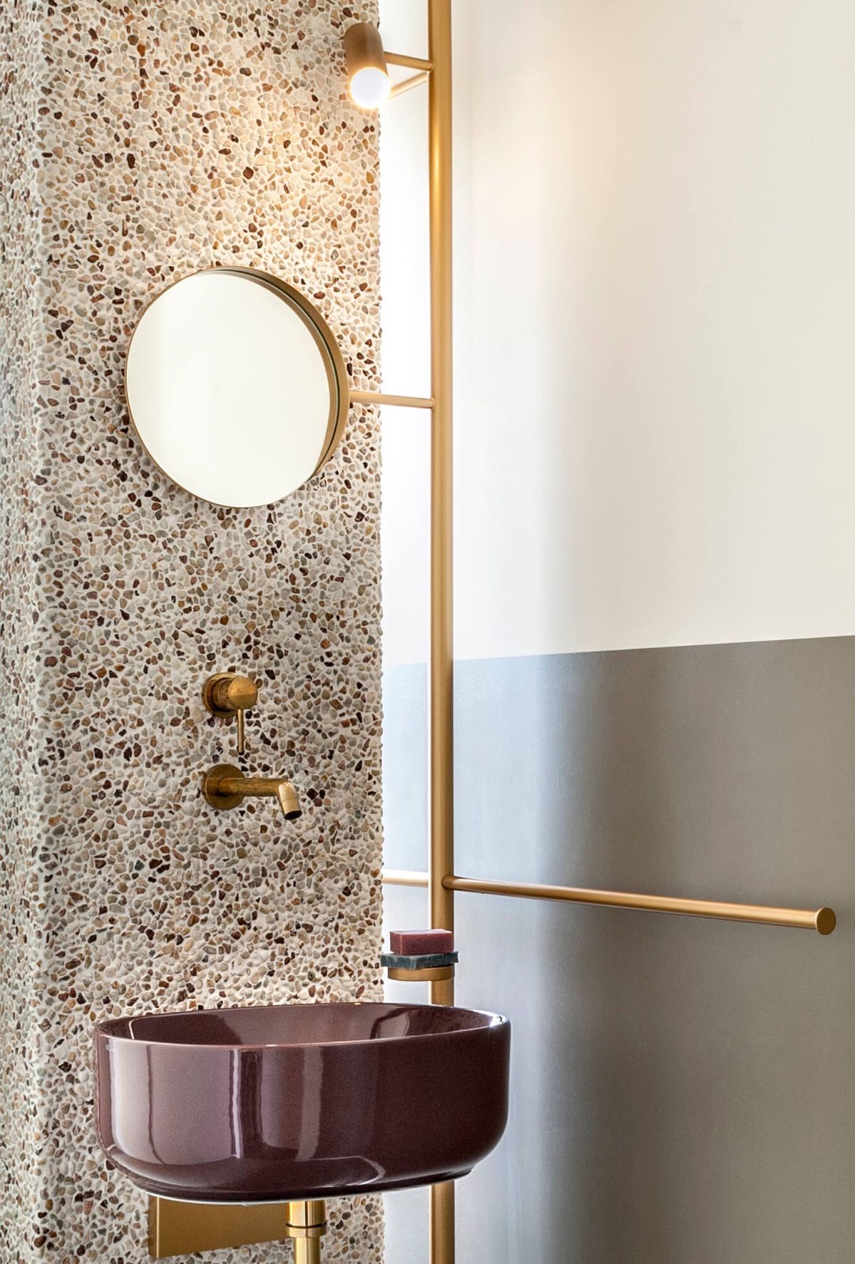 Modern luxury bathroom with a deep reddish-brown round vessel sink, a brass integrated vertical pole with mirror and towel rail, and a light-toned terrazzo/aggregate textured wall.