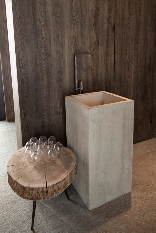 Modern industrial bathroom featuring a light grey concrete square pedestal sink, a wall-mounted chrome faucet, and a unique raw wood slice side table, set against dark wood paneling.