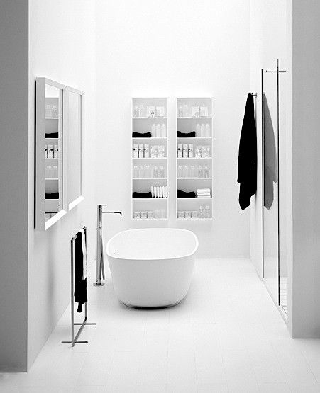 A pristine white minimalist bathroom featuring a central freestanding oval bathtub. The space has clean white walls, open wall-mounted shelves with neatly organized bath products, and a sleek freestanding faucet, creating a serene and modern spa-like ambiance.