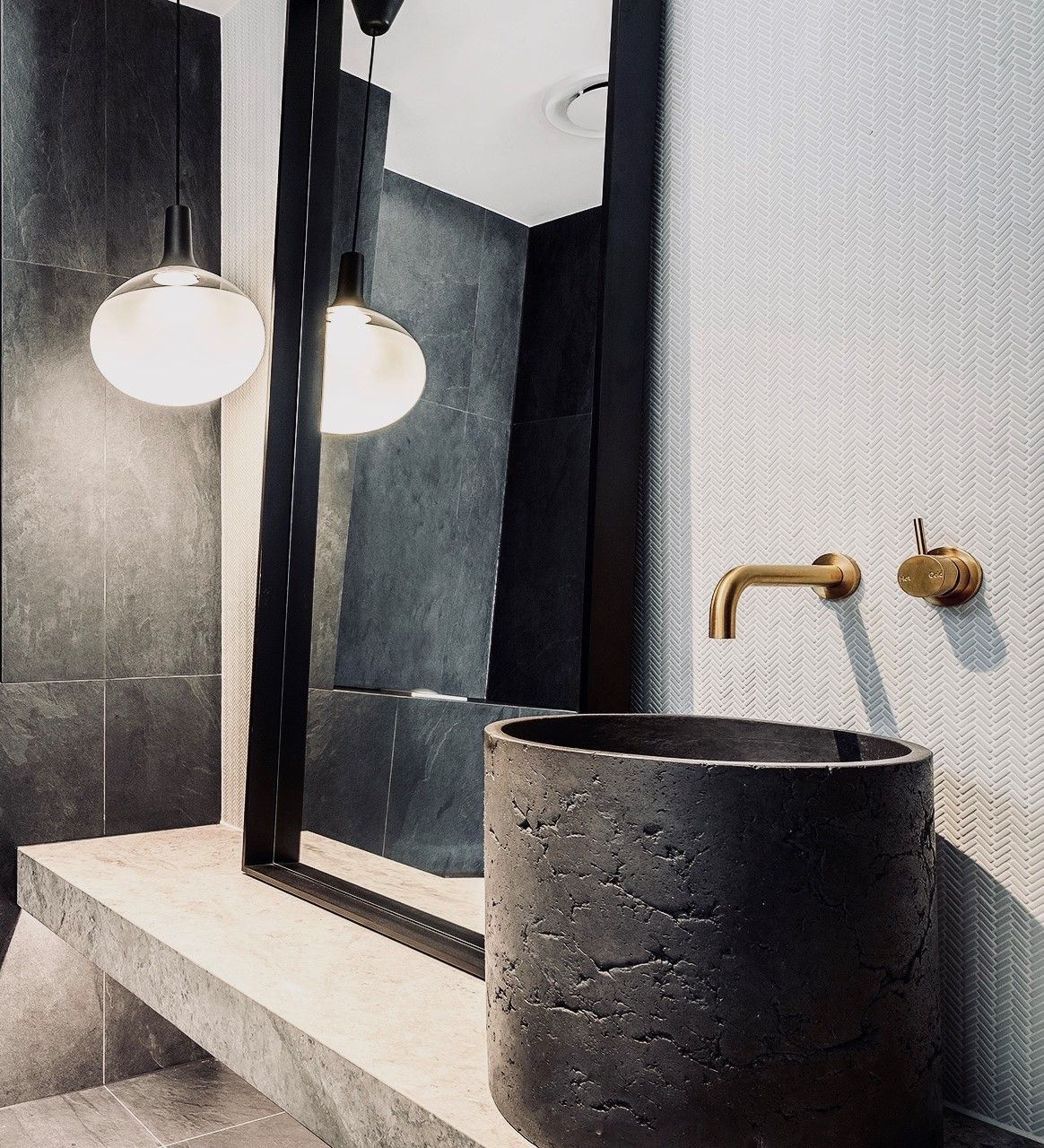 Modern luxury bathroom featuring a black rugged textured vessel sink, a brass wall-mounted faucet, and a light grey 3D geometric textured wall, with spherical pendant lights.