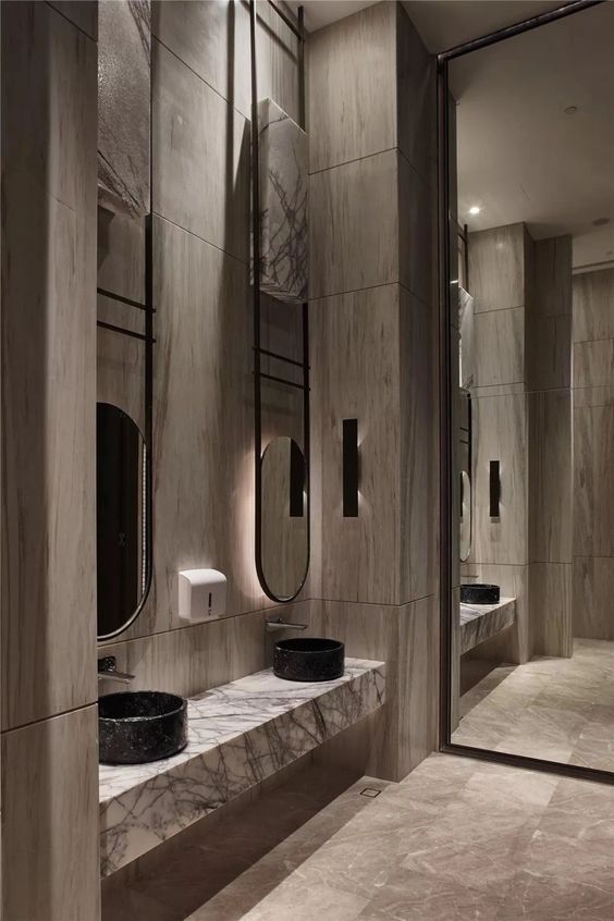 Luxurious modern public restroom with light grey marble walls and floating vanities, black round vessel sinks, slender black-framed oval mirrors, and vertical light strips.