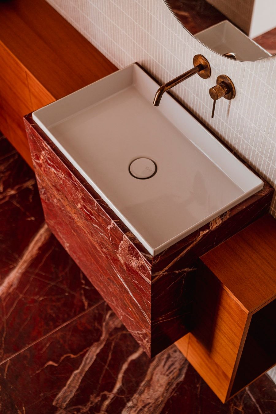 A white rectangular ceramic vessel sink sitting on a luxurious red marble vanity with a wall-mounted brass faucet and warm wood accents.