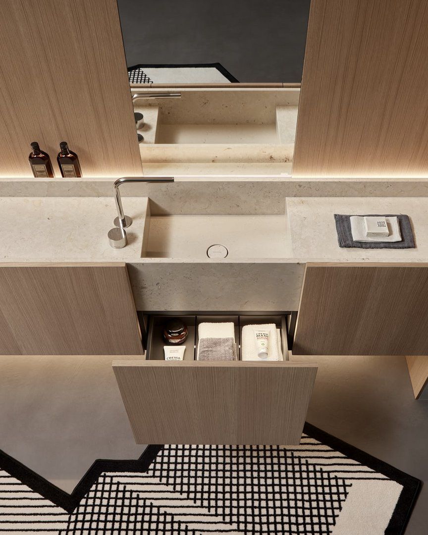 A minimalist modern bathroom featuring a light wood floating bathroom vanity with a rectangular integrated sink. The vanity has pull-out drawers, hidden LED lighting, and is complemented by a large frameless mirror and a black and white geometric patterned rug.
