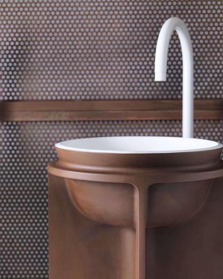 A close-up of a modern, reddish-brown freestanding washbasin with a minimalist white faucet and a white integrated basin, set against a perforated bronze-toned metal wall. The design is sleek and contemporary.