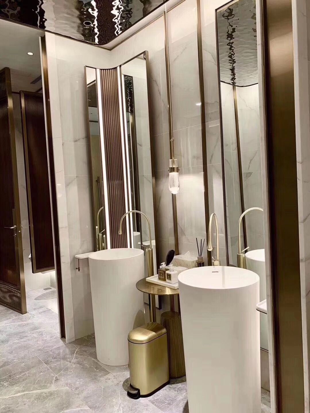 A luxurious, modern bathroom featuring a symmetrical design with two pure white freestanding cylindrical pedestal sinks. Each sink is paired with a golden curved faucet, set against mirrored walls with vertical golden accents, under a reflective ceiling, creating a high-end hotel-style ambiance.