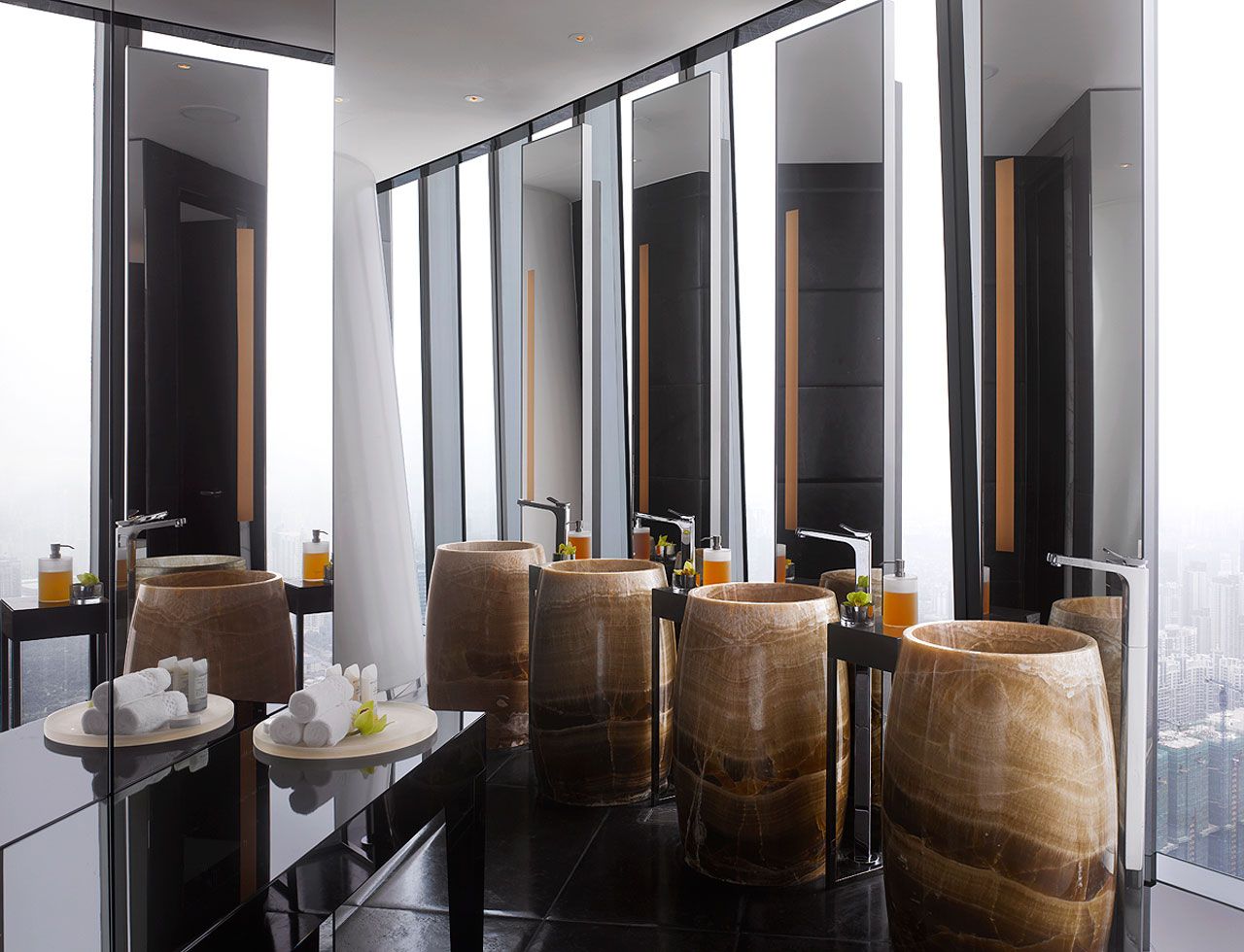 A luxurious high-rise bathroom with four cylindrical natural stone vessel sinks, likely made of translucent onyx, placed on a dark reflective vanity. The space features large floor-to-ceiling mirrored walls reflecting a city skyline, creating a grand and modern spa-like ambiance.
