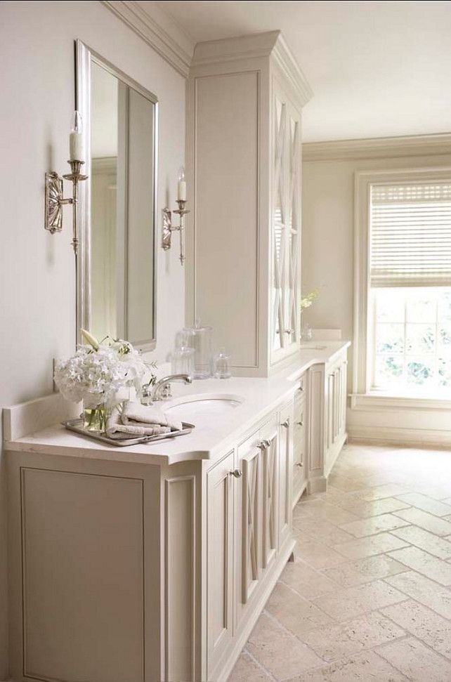 An elegant, classic bathroom featuring a long, light-colored traditional bathroom vanity with an integrated sink, polished chrome faucet, and a tall central linen tower. The walls are adorned with classic sconces and mirrors, and the floor has herringbone tile.